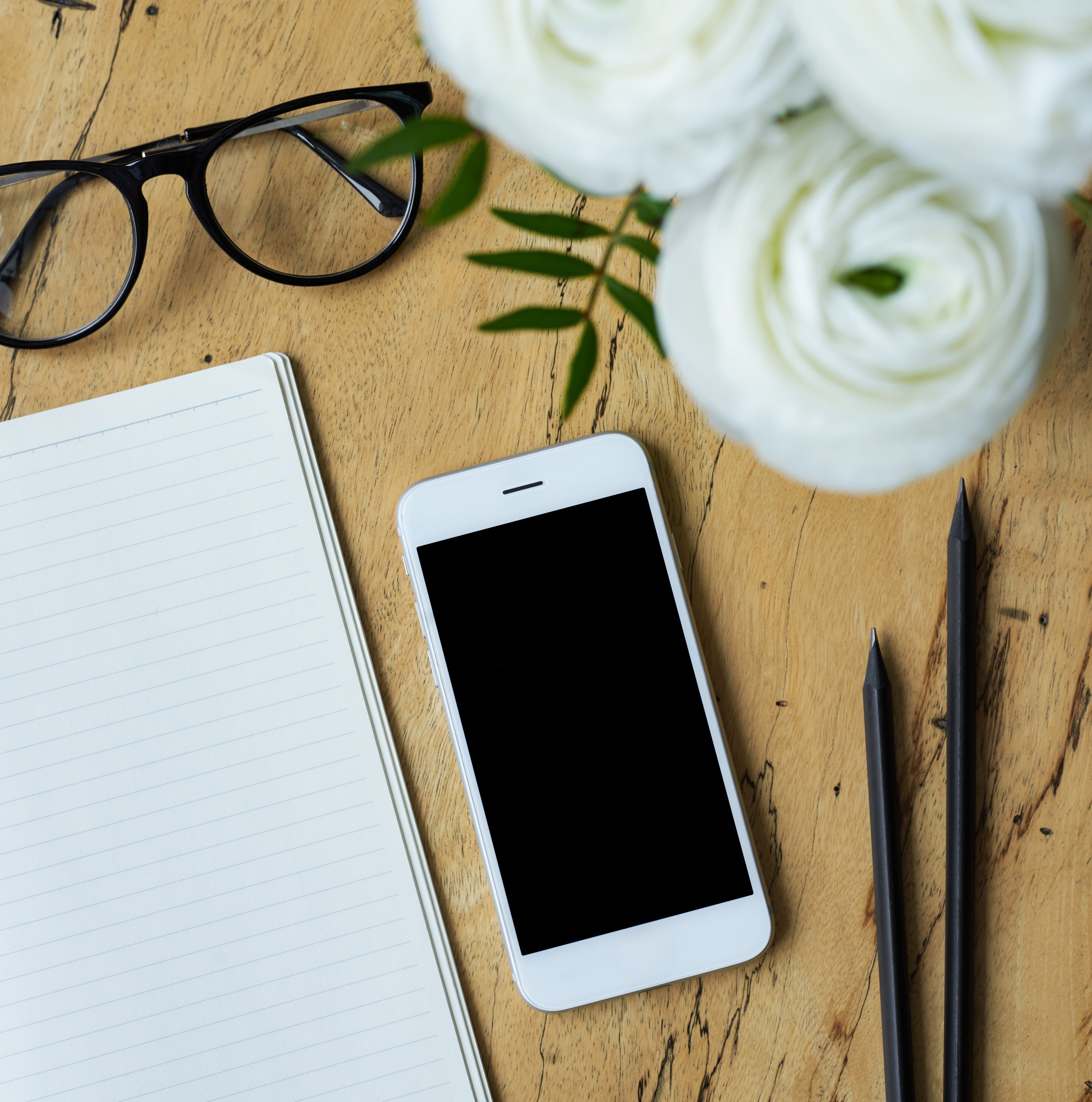 Smartphone, glasses, notebook, and pen on a wooden surface with white flowers.