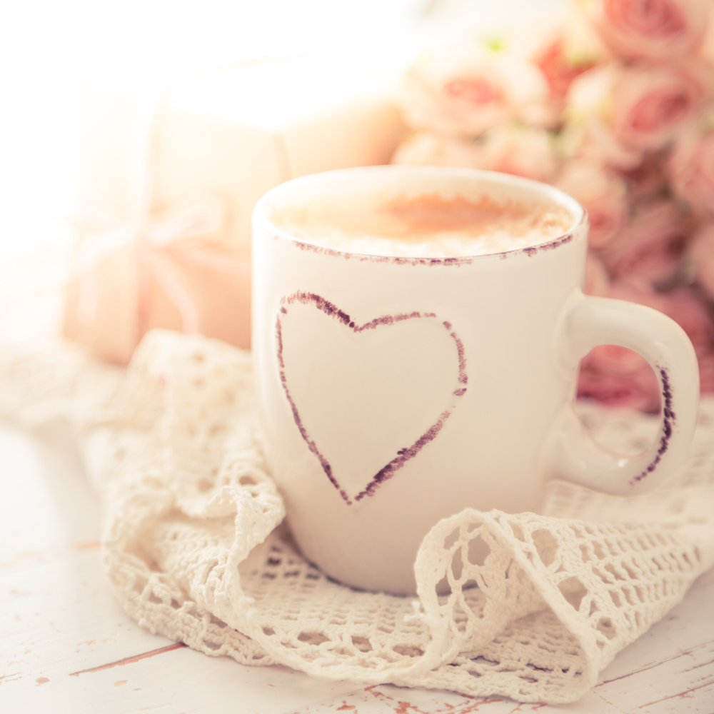 White mug with a heart design on a lace doily, blurred flowers in the background