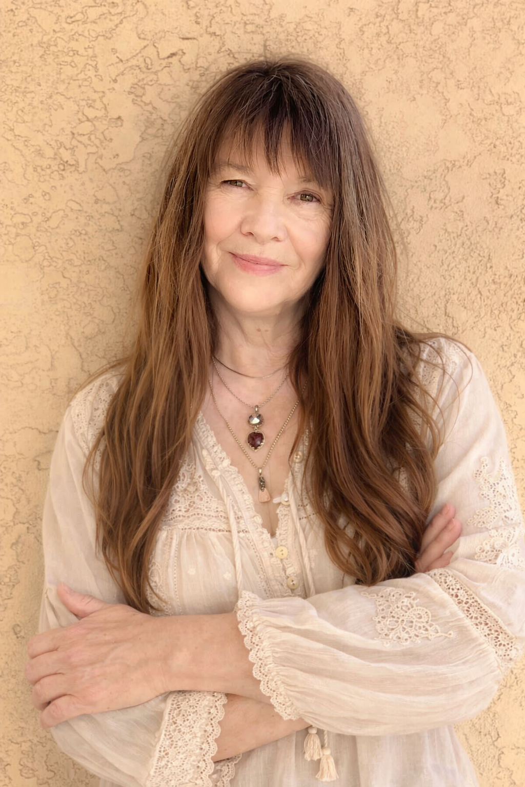 Woman with long brown hair wearing a light blouse against a beige textured wall
