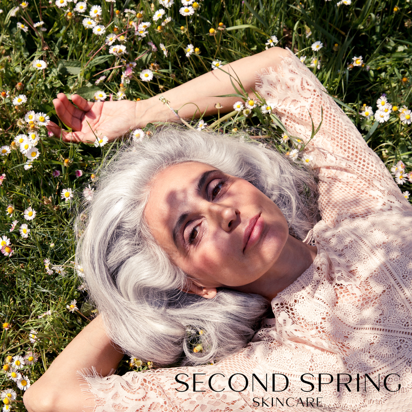 A woman with silver-gray hair lies in a sunlit field of white and yellow wildflowers, wearing a pale blush lace top. The text 'SECOND SPRING SKINCARE' appears at the bottom.