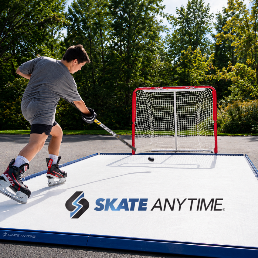 A person ice skating on a large white outdoor rink with a red hockey goal, using a hockey stick to hit a puck. The rink has the SKATE ANYTIME logo printed on the surface, with trees and pavement in the background.